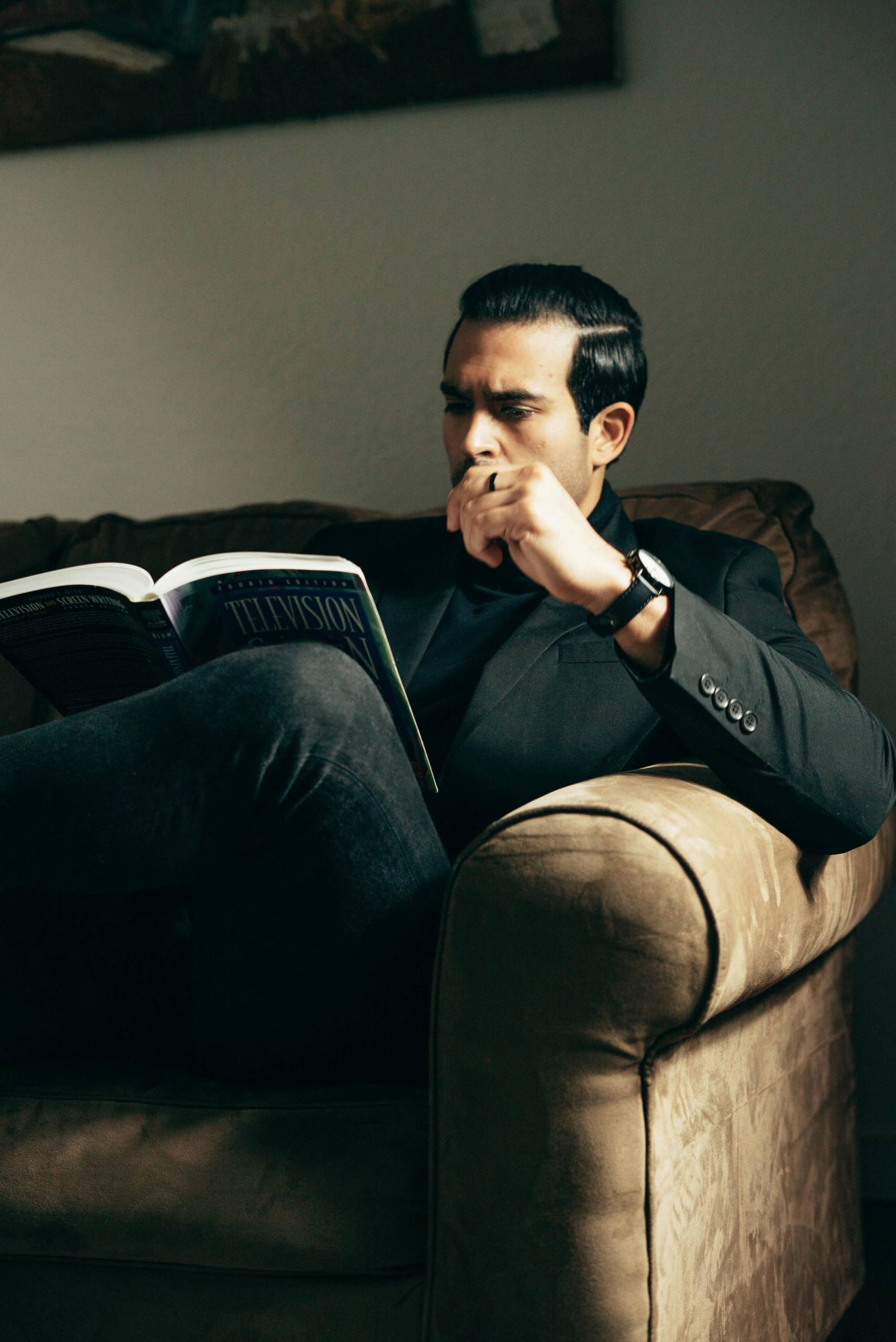 Man sitting on a couch, reading a book and thoughtfully resting his hand on his chin.